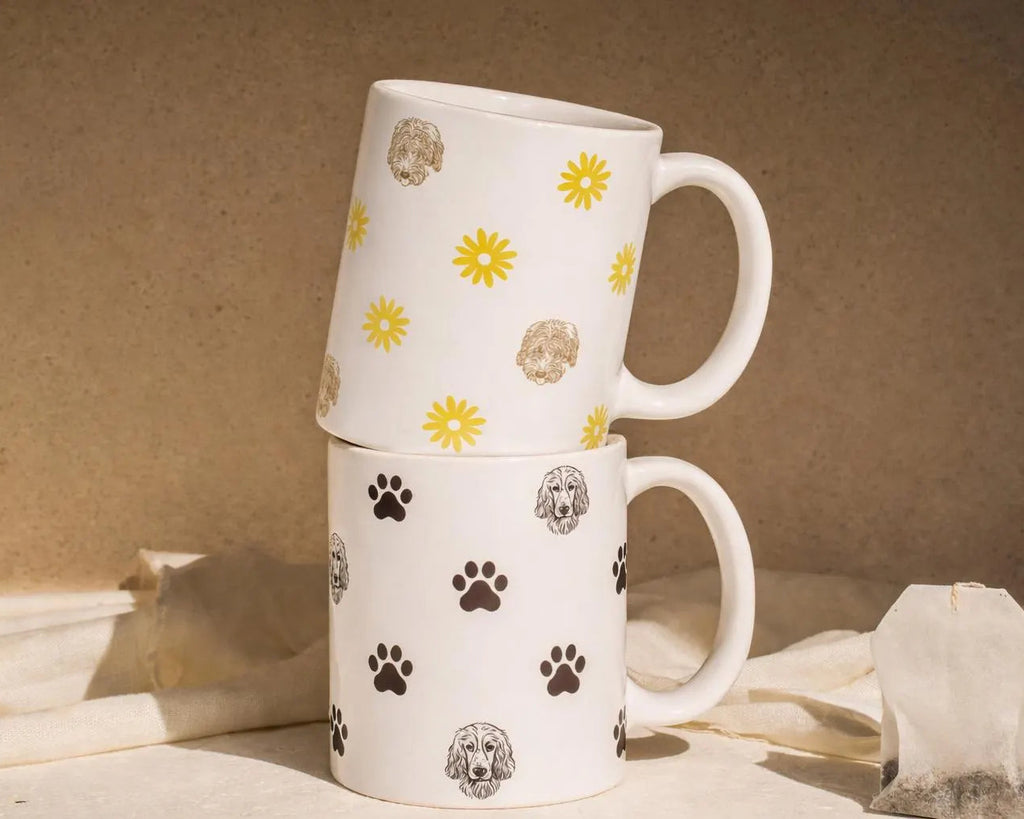 Two white mugs with floral and paw print patterns stacked on a neutral background