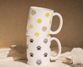 Two white mugs with floral and paw print patterns stacked on a neutral background