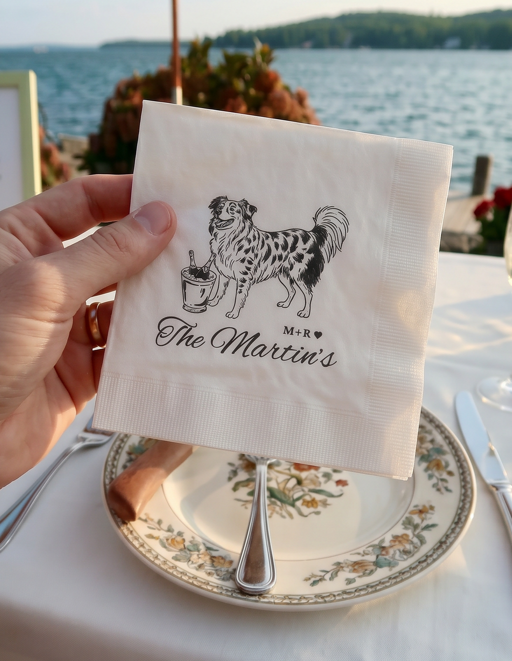Napkin with a dog illustration and 'The Martin's' text held over a table setting by a body of water.