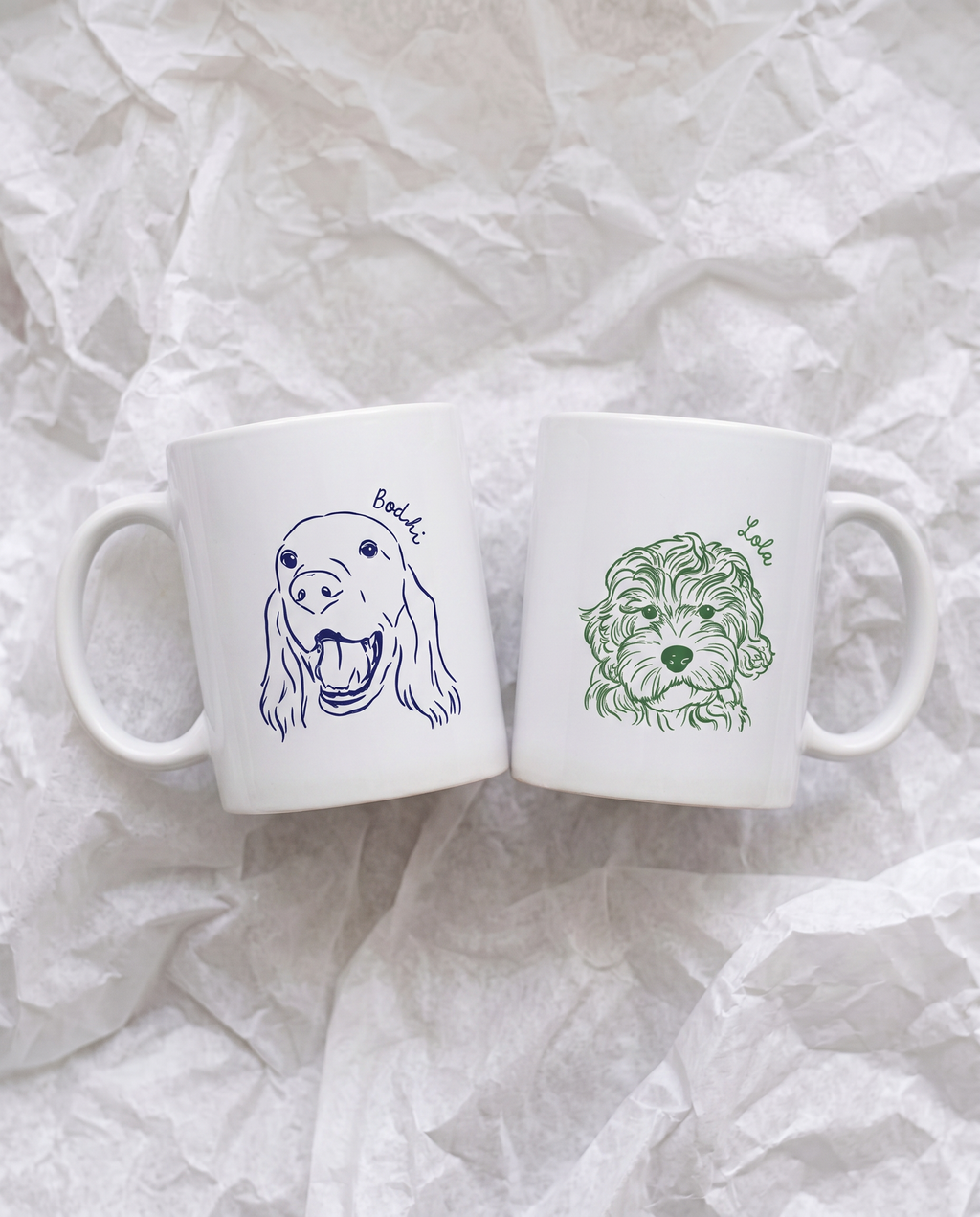Two white ceramic mugs with dog illustrations, placed on crumpled white paper. One mug shows a blue line drawing of a long-eared dog with the word 'Belgier' above it, and the other shows a green line drawing of a curly-haired dog.