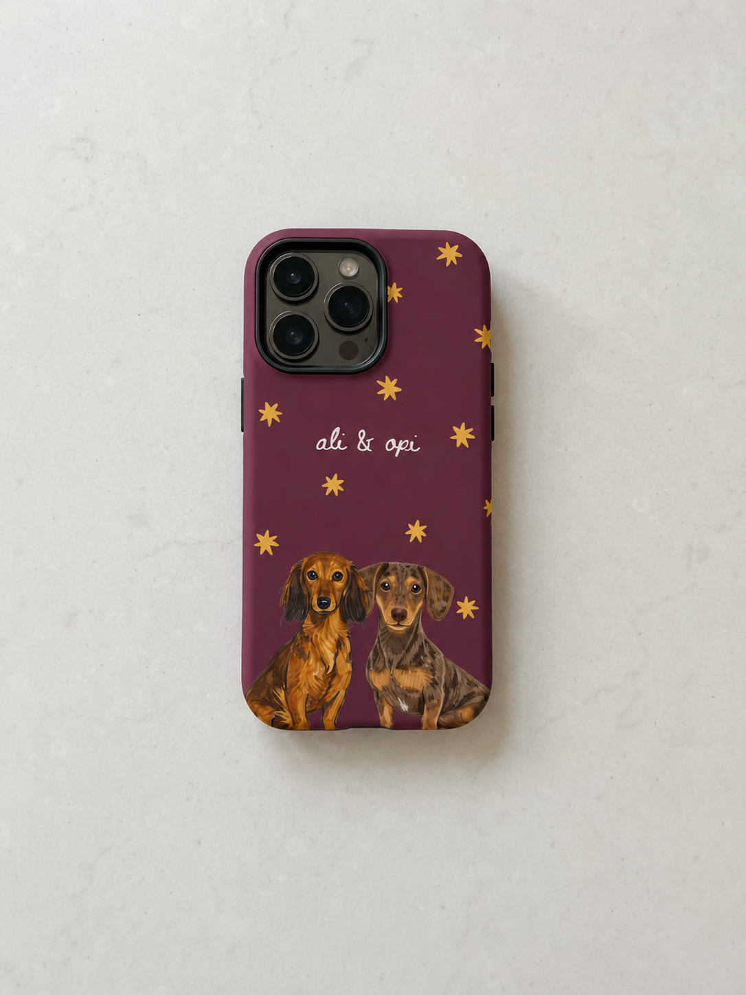 A phone case with a deep purple background, decorated with small gold stars, the text 'al. & opi', and two dachshund illustrations at the bottom, shown on a light textured surface.