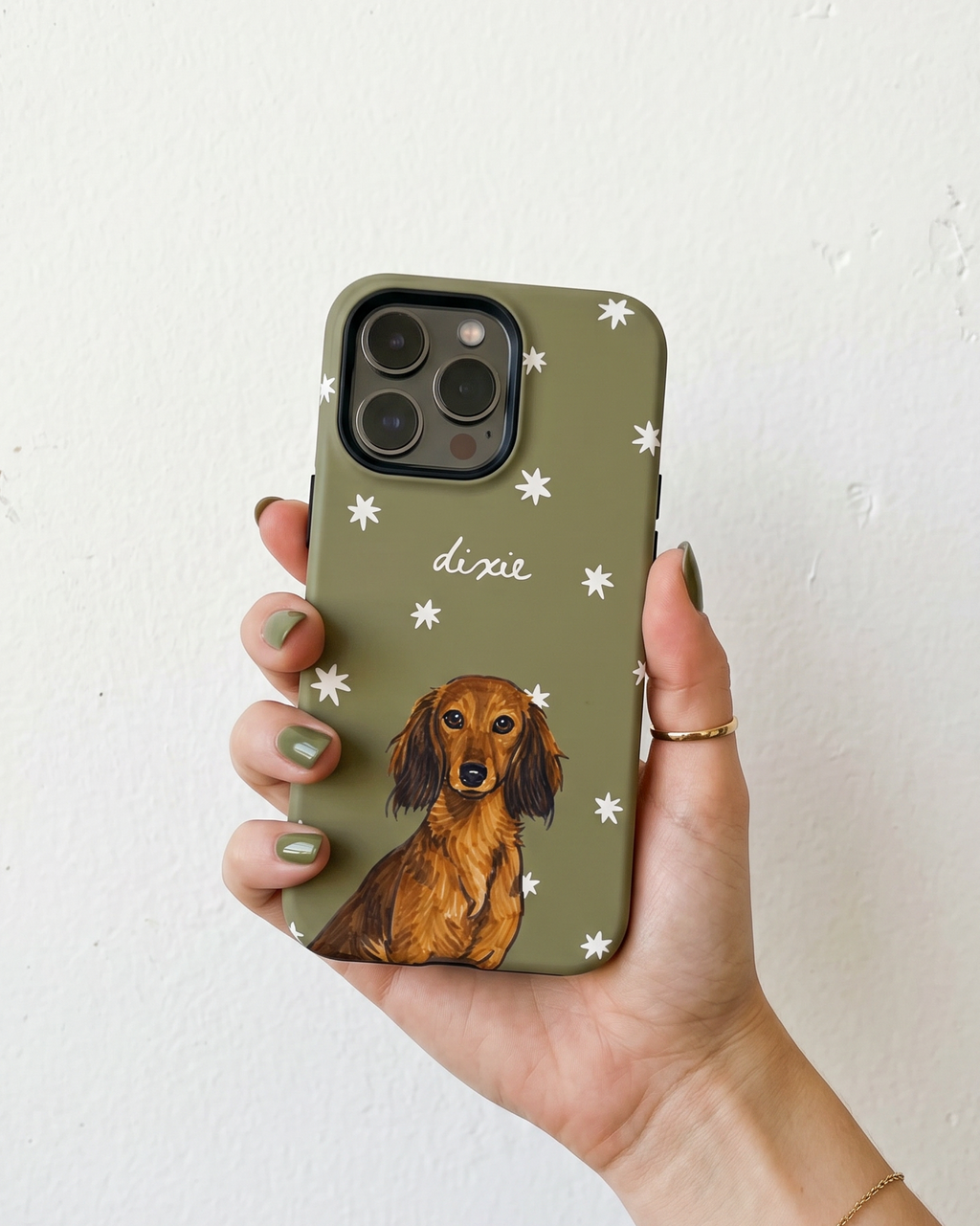 A hand holding a sage green phone case decorated with white starburst patterns, the name 'dixie' in white script, and an illustration of a brown dachshund. The hand has green nail polish and a gold ring, set against a plain white wall.