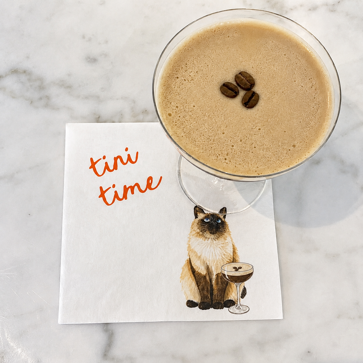 A white paper napkin printed with orange script reading 'tini time' and an illustration of a Siamese cat holding a cocktail glass, placed on a white marble surface beneath a filled margarita glass.