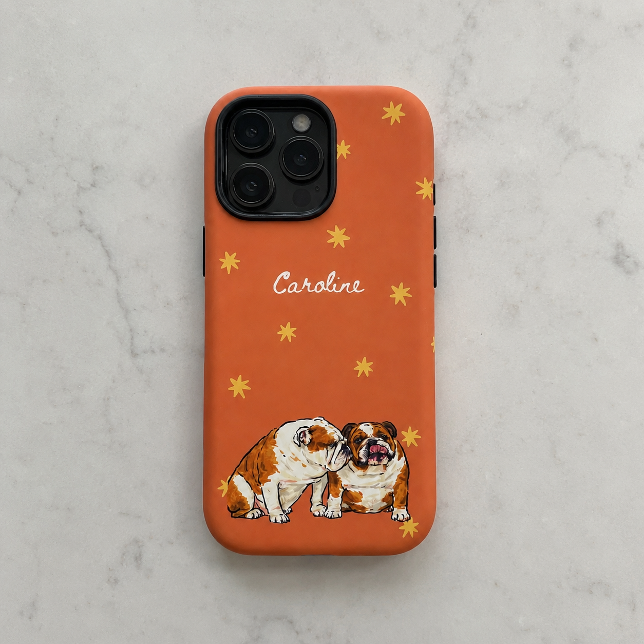 An orange phone case with a pattern of small yellow stars, the name 'Caroline' in white script, and an illustration of two bulldogs at the bottom, shown on a light gray marble surface.