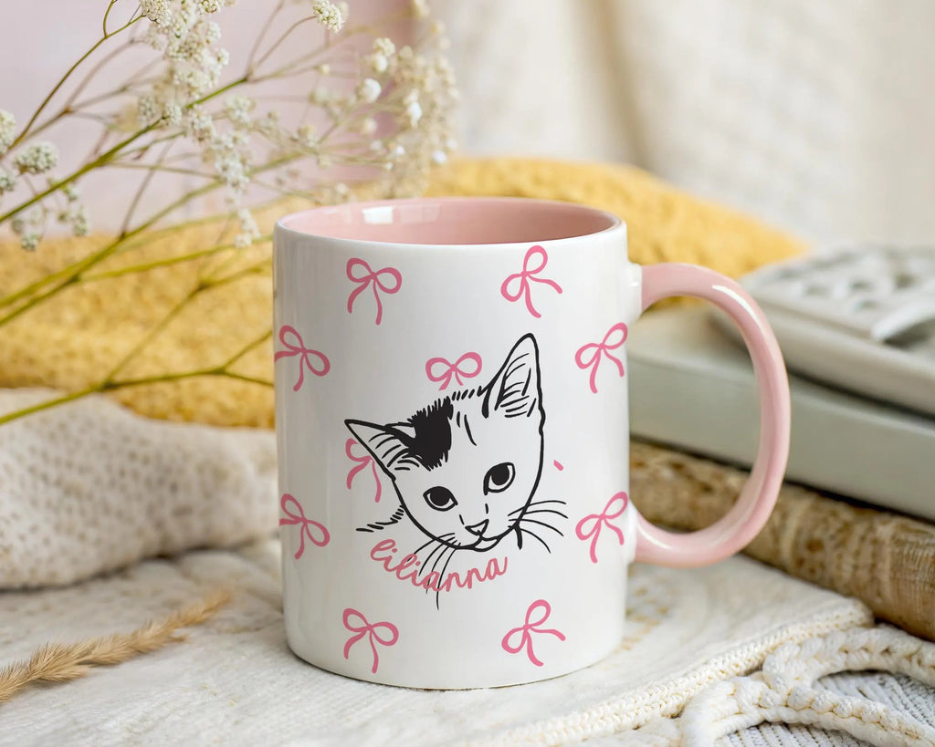 Mug with a cat illustration and pink bows on a soft surface with flowers and fabric in the background