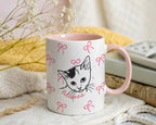Mug with a cat illustration and pink bows on a soft surface with flowers and fabric in the background