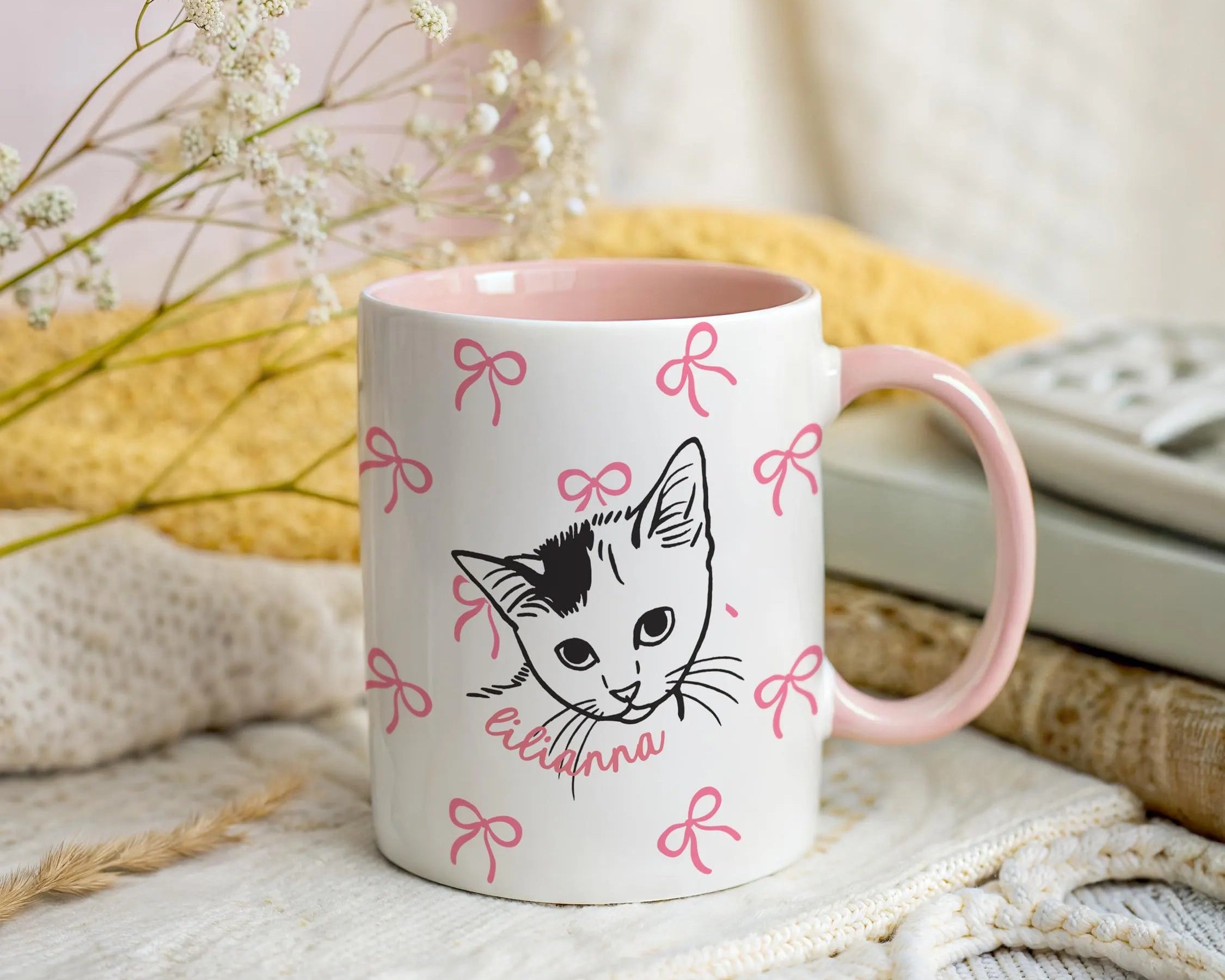 Mug with a cat illustration and pink bows on a soft surface with flowers and fabric in the background