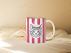 Mug with a cat illustration and name on a striped pattern, placed on a soft surface.