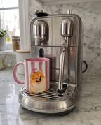 A stainless steel espresso machine on a marble countertop with a pink and white striped ceramic mug featuring a Pomeranian dog illustration placed under the steam wand.