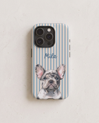A personalized phone case featuring a French Bulldog illustration with the name 'Mila' in blue script, set against a light blue and white striped background, displayed on a light beige textured surface.