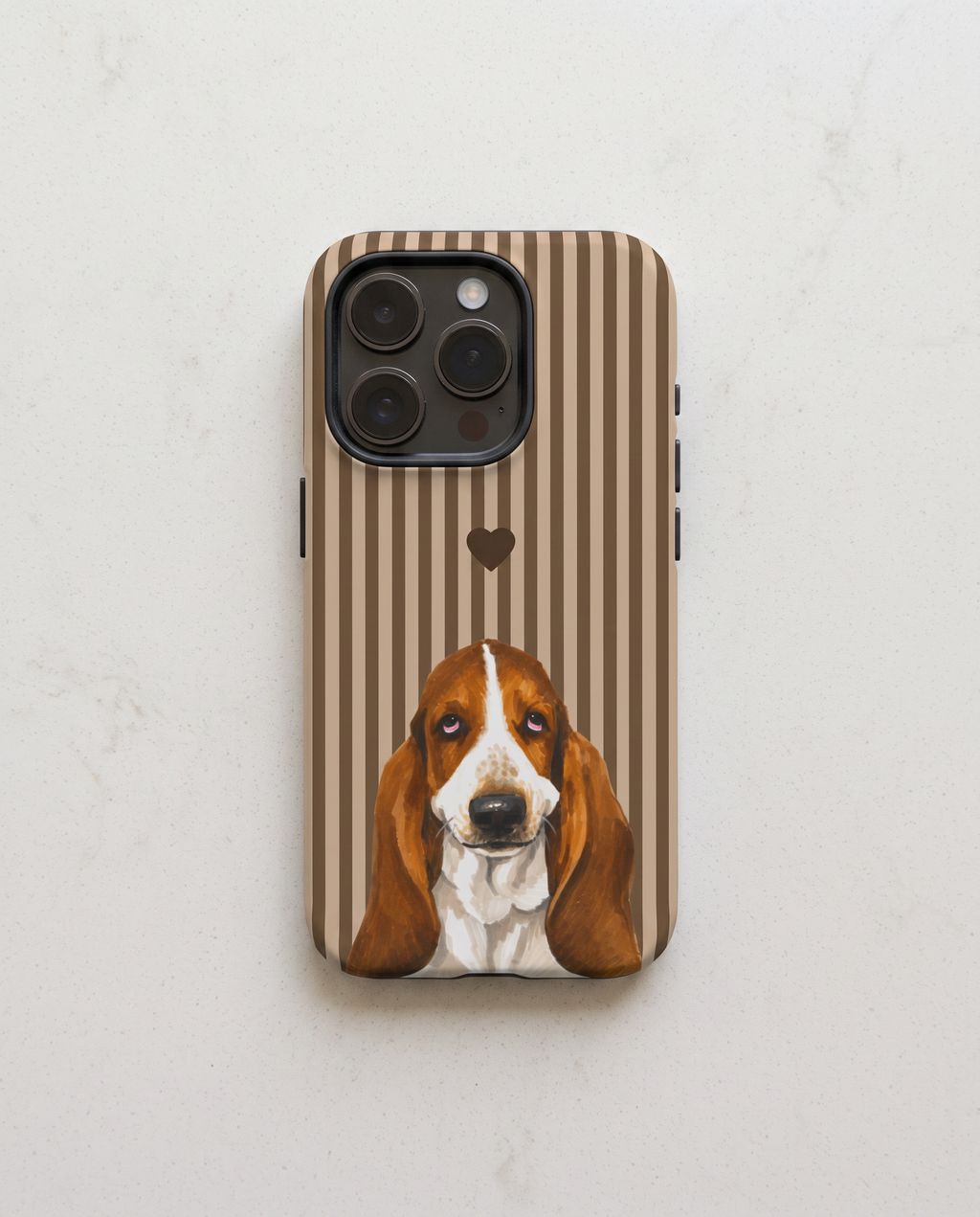 A phone case featuring a brown and white striped background with a painted Basset Hound illustration and a small heart, shown on a light marble surface.