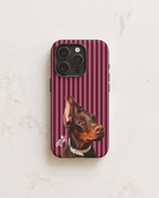 A phone case featuring a portrait of a Doberman dog on a burgundy and pink striped background, shown on a light marble surface.