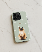 A sage green phone case featuring a watercolor illustration of a Siamese cat and the name 'kitty' in brown, shown on a light marble background.