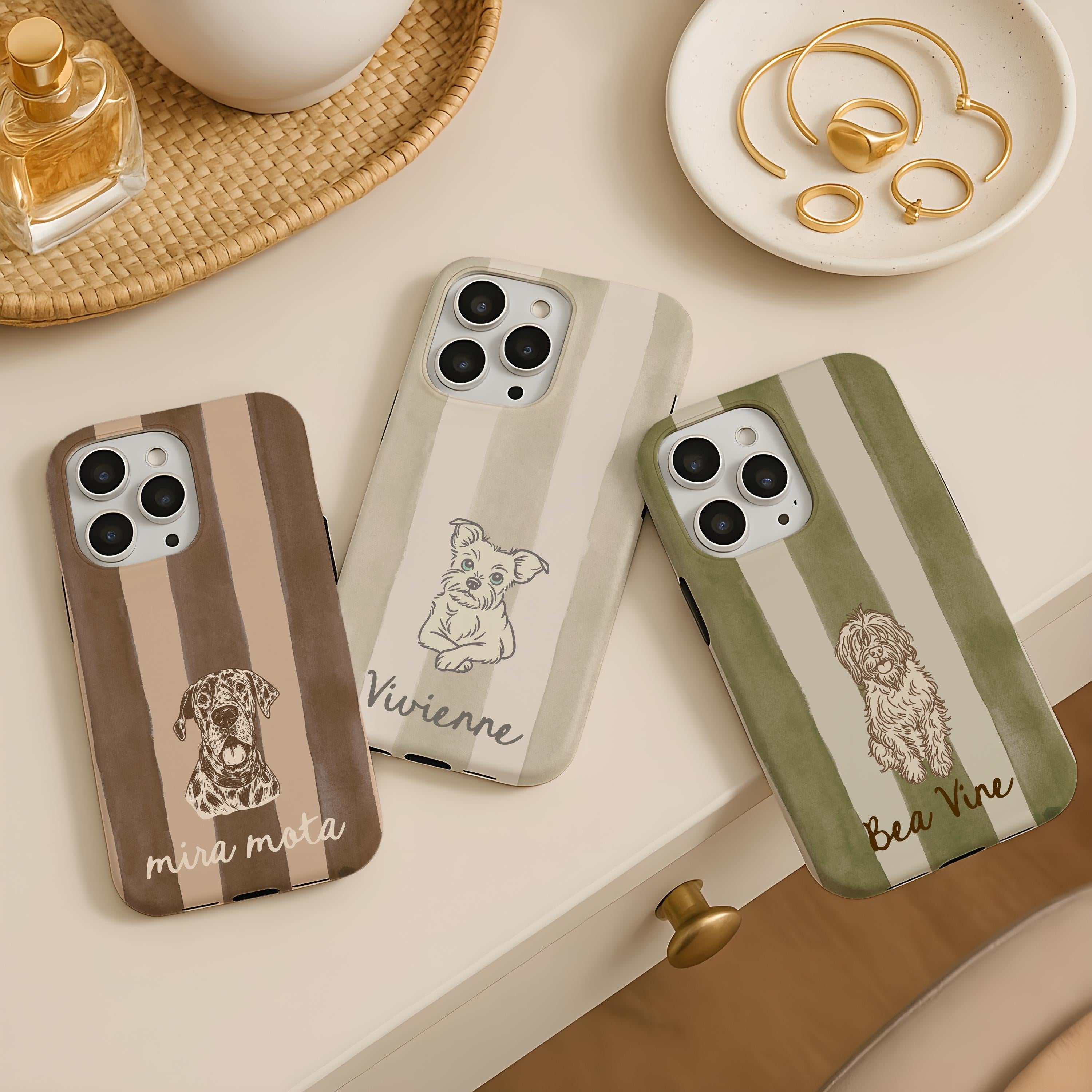Three custom striped phone cases with dog illustrations and names on a table with a perfume bottle and jewelry.