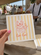 A hand with pink nail polish holds a square paper napkin featuring pale yellow vertical stripes and a coral-red line drawing of a dog holding a cocktail, with the text 'Cin Cin!' below. The background is a blurred outdoor bar or event setting with people and a bartender.
