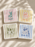 Four square paper cocktail napkins with striped backgrounds and dog-themed illustrations, arranged in a 2x2 grid on a light beige surface with soft leaf shadows. The napkins feature pink, yellow, green, and blue stripes with drawings of dogs and text
