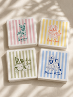 Four square paper cocktail napkins with striped backgrounds and dog-themed illustrations, arranged in a 2x2 grid on a light beige surface with soft leaf shadows. The napkins feature pink, yellow, green, and blue stripes with drawings of dogs and text