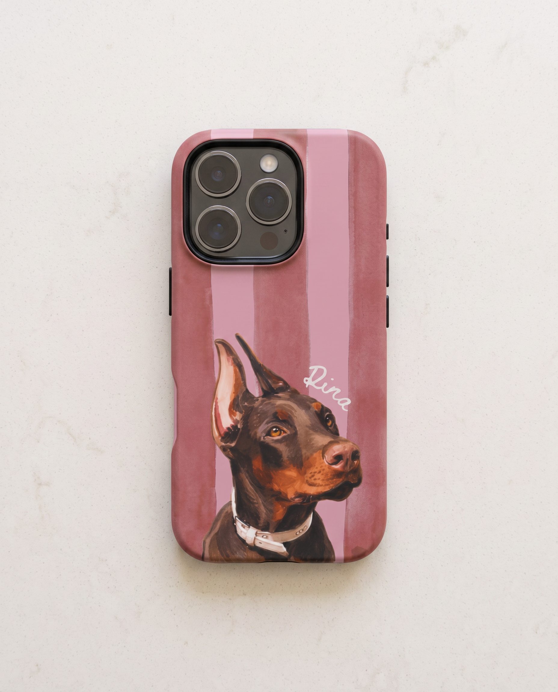 A protective phone case with a vertical dusty pink and mauve stripe pattern, featuring a custom painted portrait of a Doberman dog named 'Rina' on the back, displayed on a light marble surface.