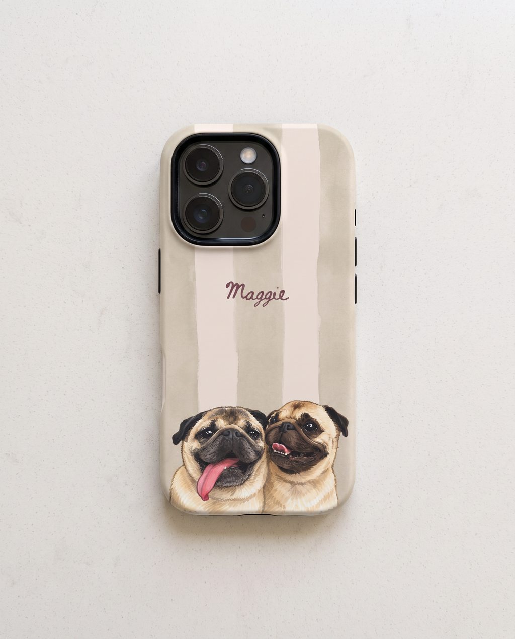 A personalized phone case featuring two illustrated pug dogs with their tongues out, set against a vertical beige and cream stripe pattern with the name 'Maggie' written in brown cursive. The case is shown on a light gray textured surface.