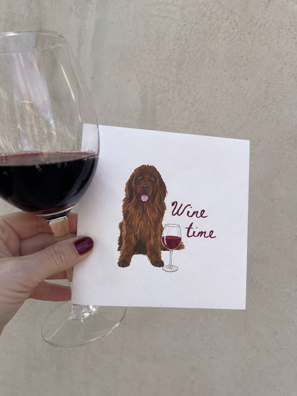 A hand holds a white greeting card featuring an illustration of a brown long-haired dog next to a wine glass and the words 'Wine time'. A real wine glass filled with red wine is partially visible on the left against a neutral beige wall.