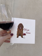 A hand holds a white greeting card featuring an illustration of a brown long-haired dog next to a wine glass and the words 'Wine time'. A real wine glass filled with red wine is partially visible on the left against a neutral beige wall.